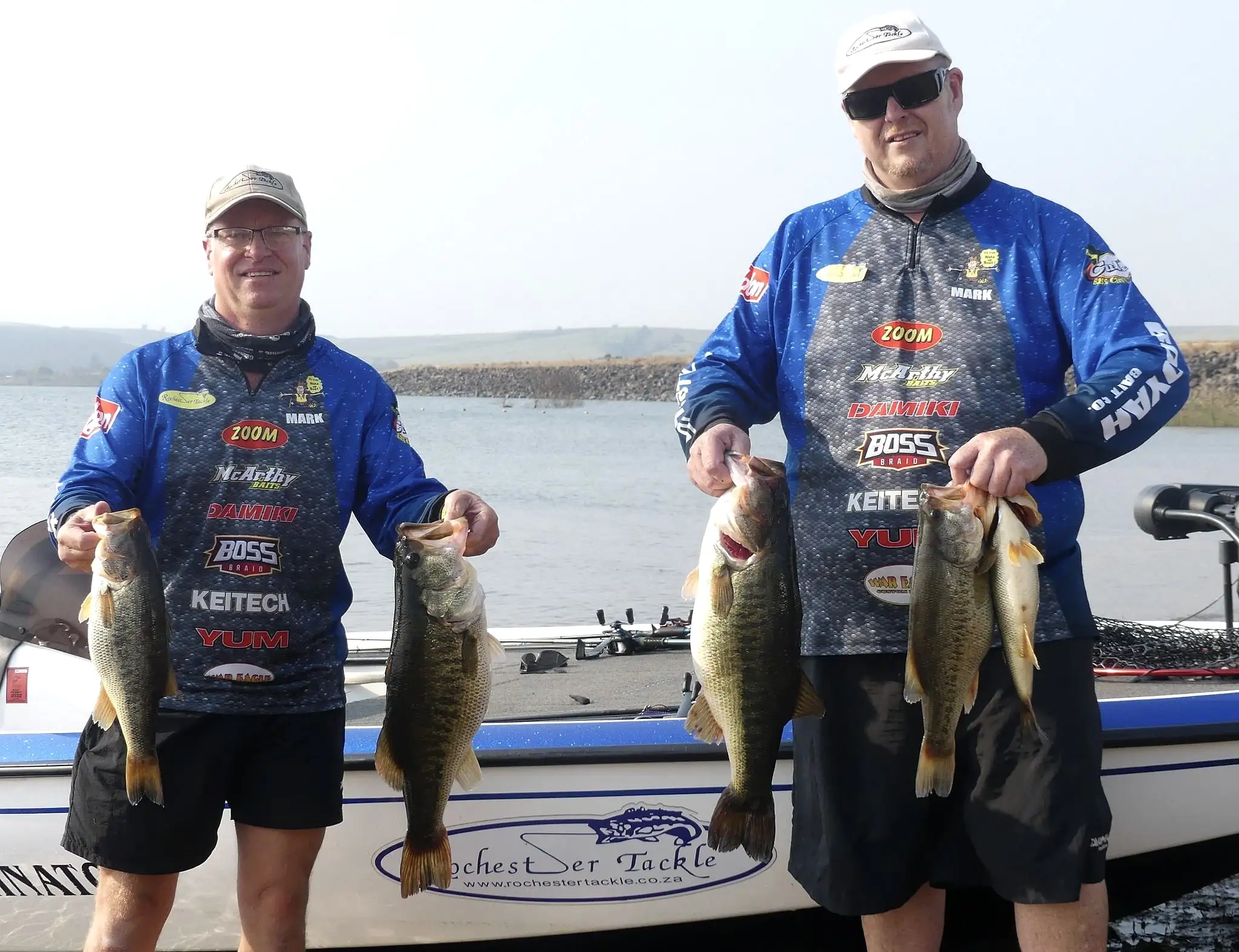 Two anglers in Rochester jerseys holding four bass beside the Rochester Tackle boat at Albert Falls