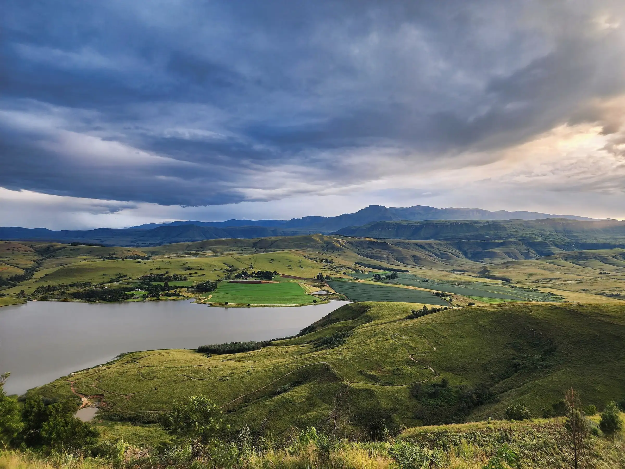 A dam among green hills with the Drakensberg under a heavy sky