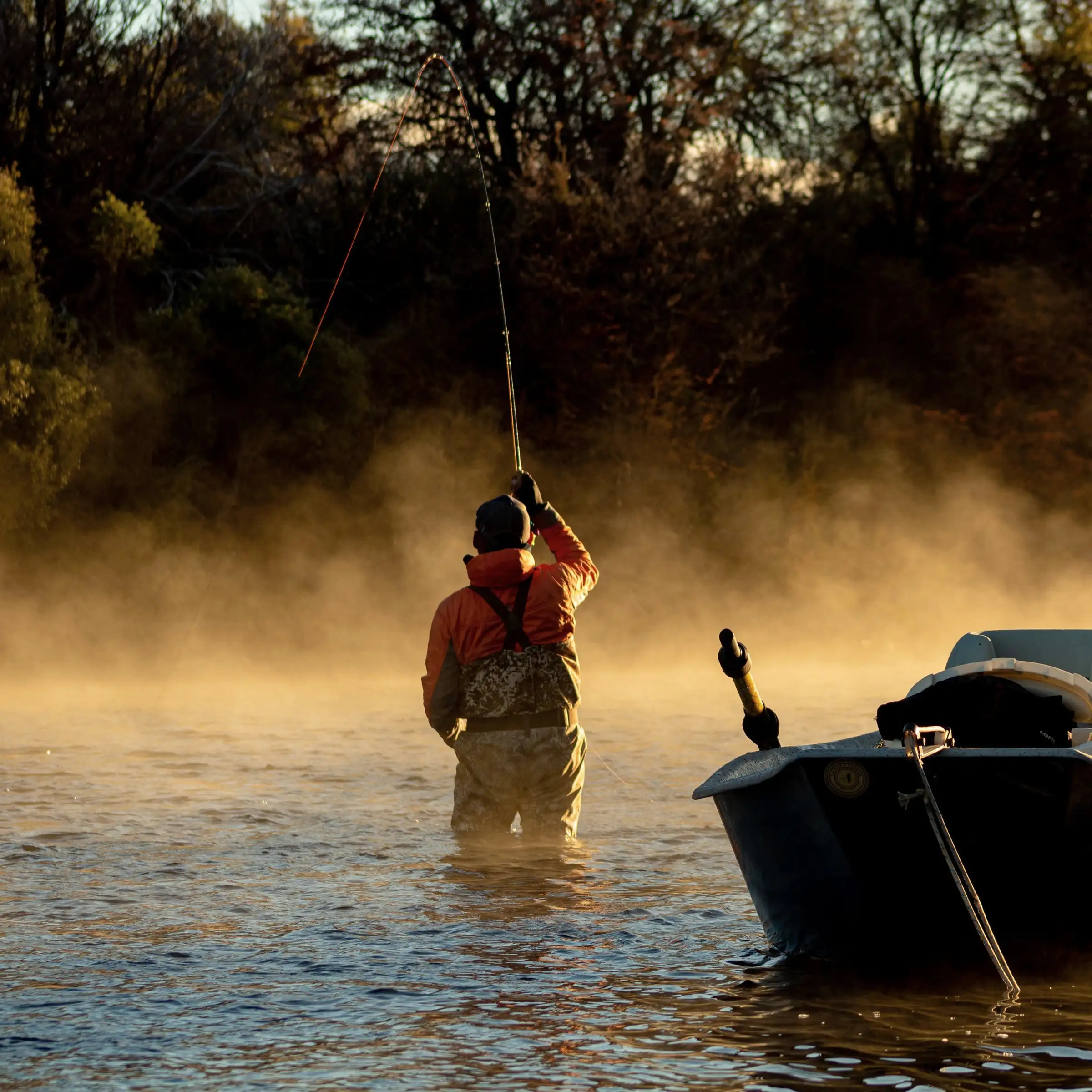 A fly angler casting in a misty river with a drift boat beside him