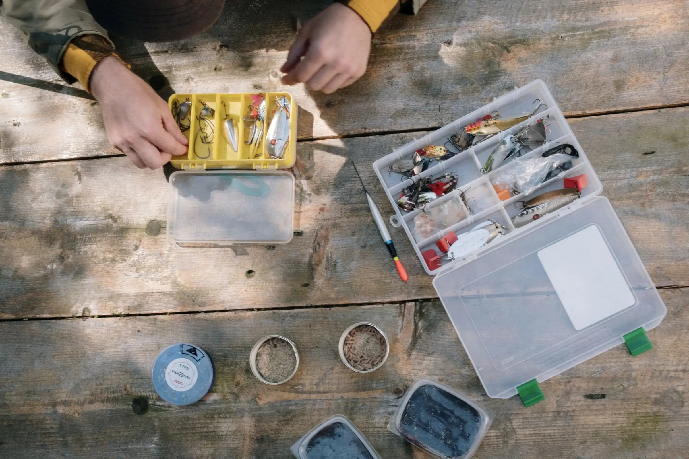 Hands sorting lures in open tackle boxes on a wooden table