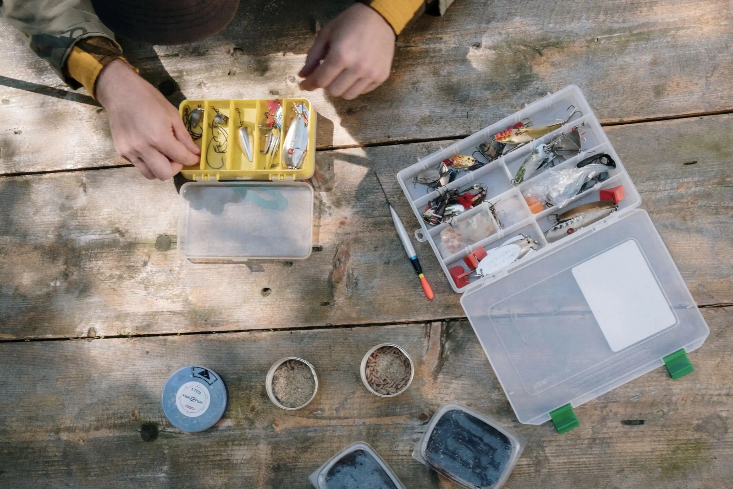 Hands sorting lures in open tackle boxes on a wooden table