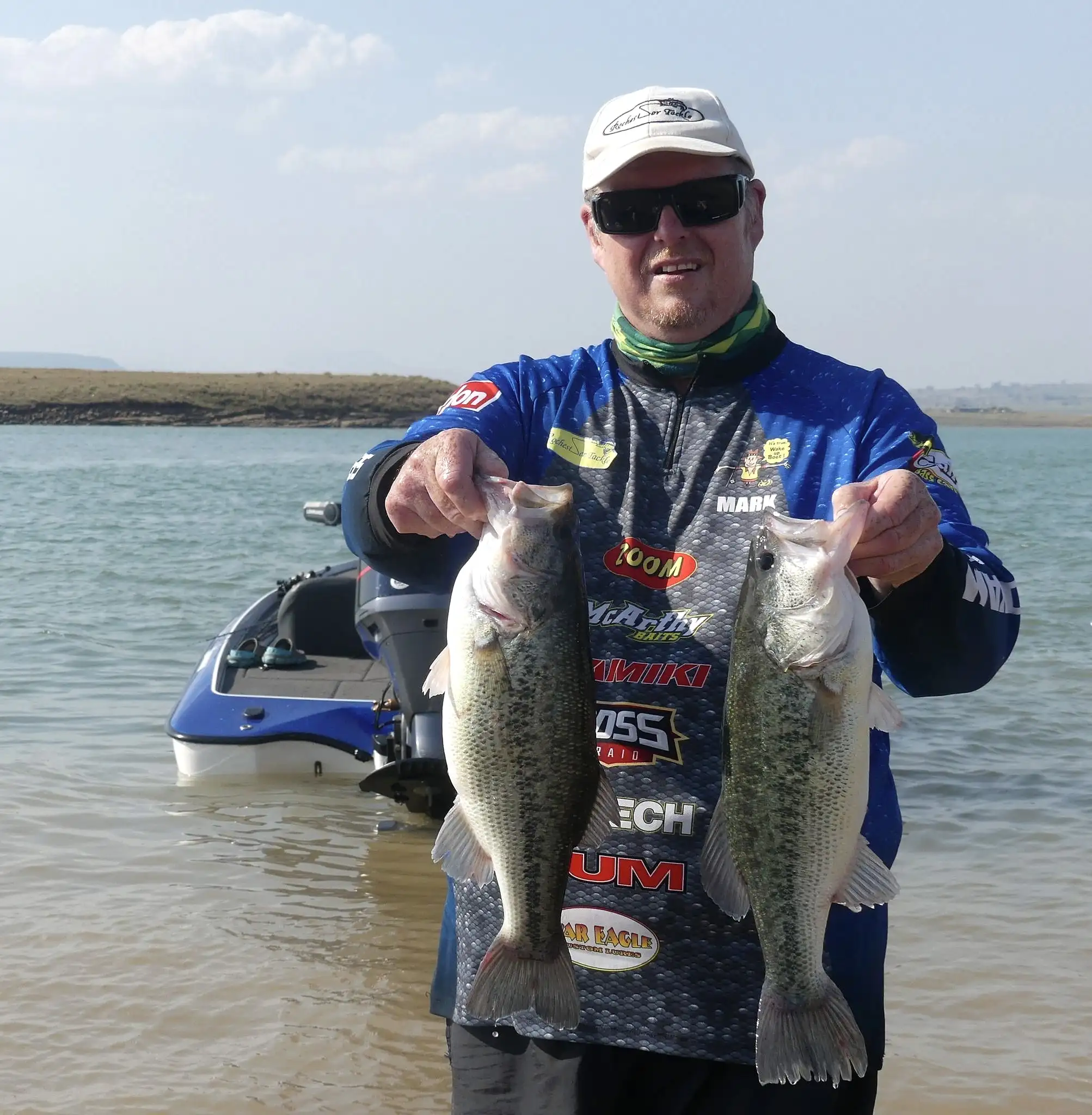 An angler in a tournament shirt holding two bass at the water's edge